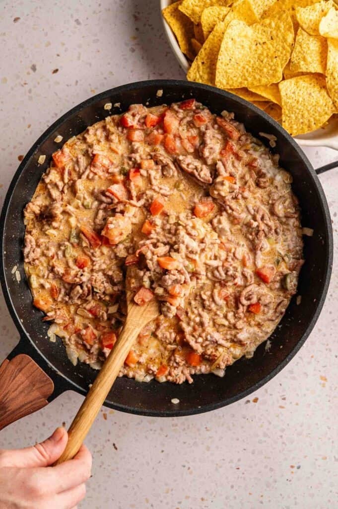 A hand stirring a creamy mixture of ground beef, diced tomatoes, and onions for Dorito Casserole in a black skillet with a wooden spoon, with a bowl of nacho cheese Doritos in the background.