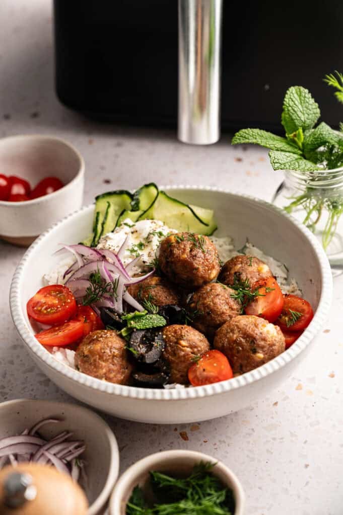 A bowl of seven Air Fryer Lamb Meatballs served over white rice with tzatziki, sliced cucumbers, red onions, cherry tomatoes, and black olives, garnished with fresh dill and mint, alongside small bowls of extra tomatoes, onions, herbs, and a sprig of mint in a glass jar.