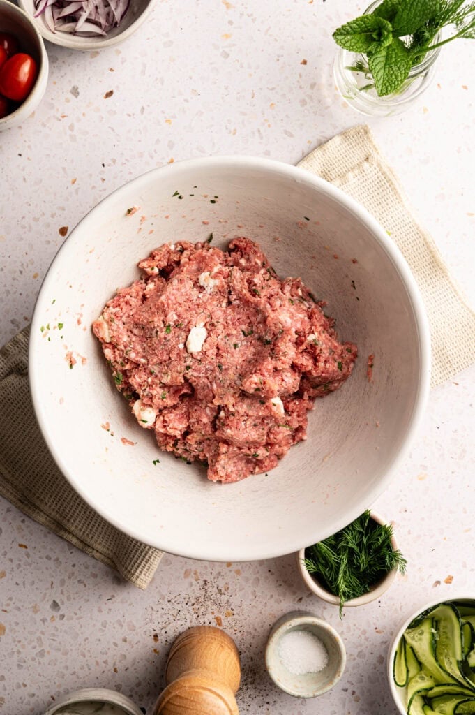 A bowl of raw Air Fryer Lamb Meatball mixture combined with herbs and feta cheese, surrounded by prep bowls of cherry tomatoes, sliced red onions, fresh dill, sliced cucumbers, salt, and a sprig of mint in a glass jar.