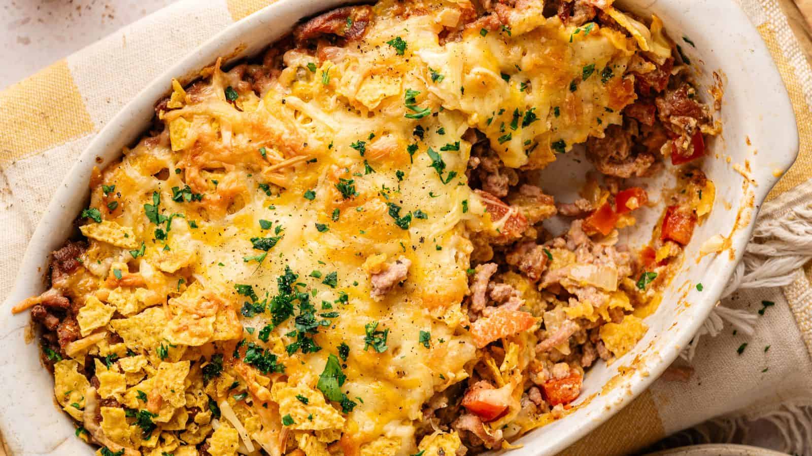 A baked casserole topped with melted cheese and crushed tortilla chips, with a portion removed to show layers of ground meat, vegetables, and more chips inside.
