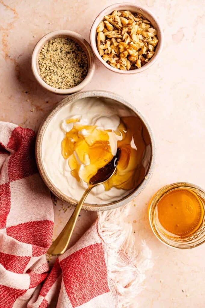 Ninja Creami frozen yogurt ingredients: Greek yogurt with honey being stirred by a gold spoon, hemp seeds, and chopped walnuts on a marble surface.