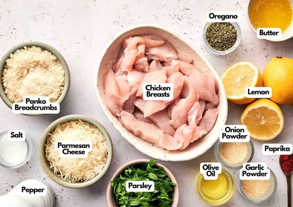 An ingredient layout for Air Fryer Parmesan Crusted Chicken featuring sliced raw chicken breasts in a white bowl, surrounded by panko breadcrumbs, shredded parmesan cheese, and fresh parsley. The spread includes small bowls of melted butter, olive oil, oregano, onion powder, garlic powder, salt, and pepper, with a spoonful of paprika and three lemon halves arranged on a speckled light pink background.
