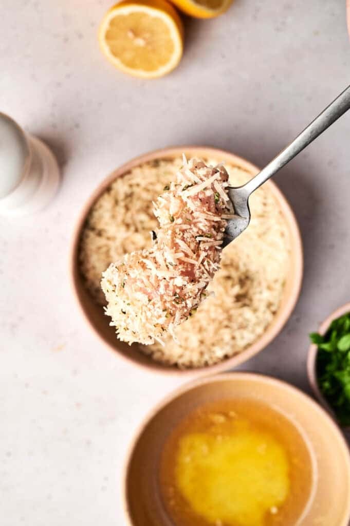 A single piece of raw chicken on a fork being coated in a Parmesan and breadcrumb mixture, held over a bowl of breading with a bowl of melted butter, fresh parsley, a white salt shaker, and lemon halves in the background.