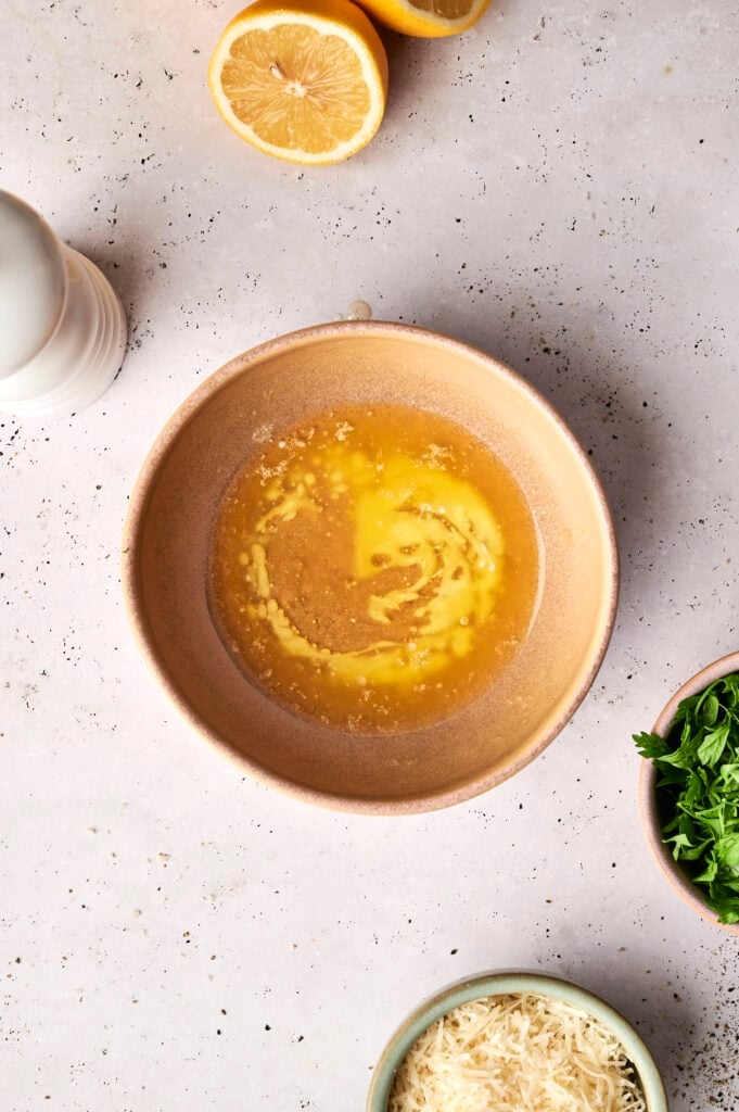 A bowl of melted butter for Air Fryer Parmesan Crusted Chicken, surrounded by background elements including two lemon halves, a white salt shaker, a small bowl of fresh parsley, and a bowl of Parmesan cheese.
