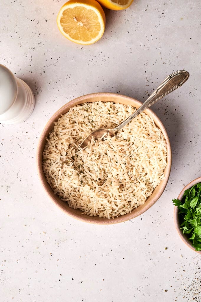 A bowl of Parmesan cheese and breadcrumb coating for Air Fryer Parmesan Crusted Chicken, with a silver spoon inside, surrounded by background elements including lemon halves, a white salt shaker, and a small bowl of fresh parsley.