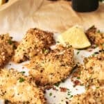Six pieces of golden-brown Air Fryer Parmesan Crusted Chicken on parchment paper, garnished with fresh chopped parsley and a lemon wedge, with a black pepper grinder in the background.