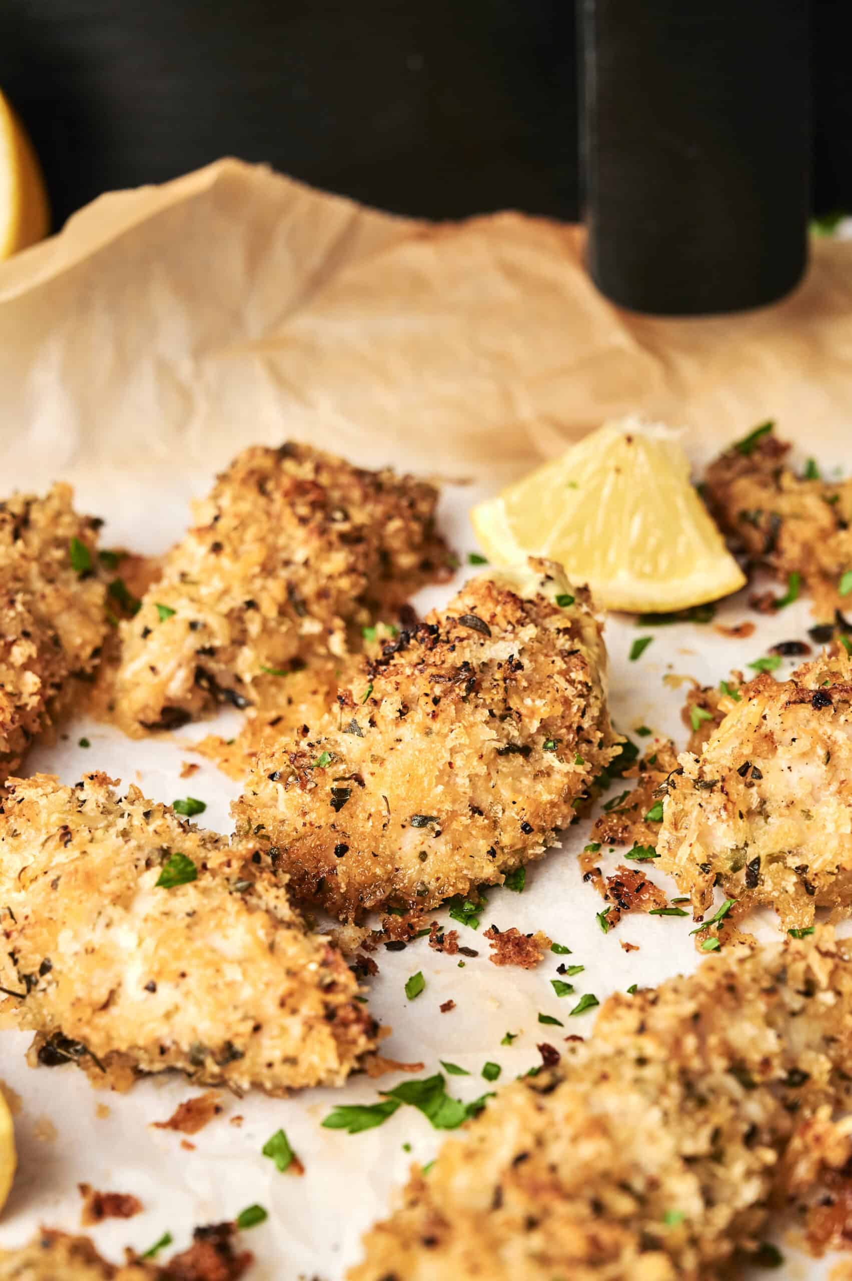 Six pieces of golden-brown Air Fryer Parmesan Crusted Chicken on parchment paper, garnished with fresh chopped parsley and a lemon wedge, with a black pepper grinder in the background.