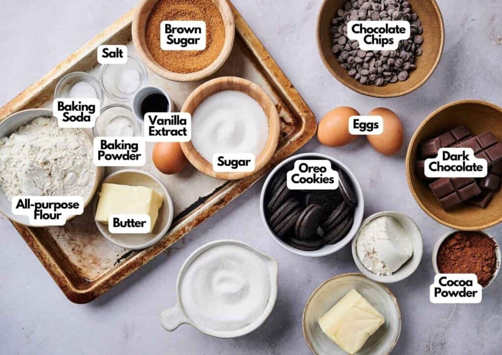 An overhead shot of ingredients for air fryer slutty brownies arranged on a rustic, worn baking sheet and a light grey surface, featuring bowls of all-purpose flour, brown sugar, white sugar, chocolate chips, cocoa powder, dark chocolate chunks, and Oreo cookies, alongside three eggs, two blocks of butter, vanilla extract, baking soda, baking powder, and salt.