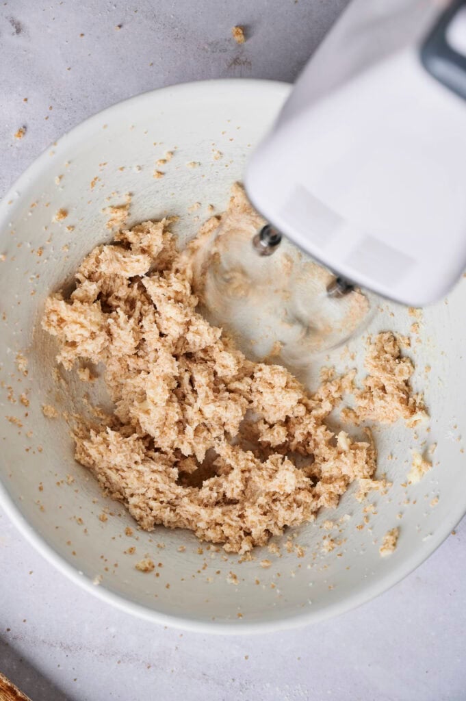 A top-down view of a single, uncut square of Air Fryer Slutty Brownies with a crinkle top, sitting in a black air fryer basket lined with white parchment paper, with a bowl of chocolate chips visible in the upper left corner.