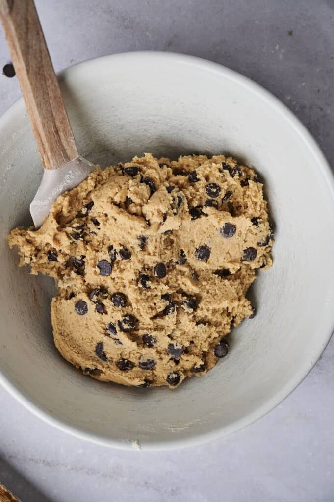 A bowl of chocolate chip cookie dough being mixed with a wooden-handled spatula, prepared as the base layer for Air Fryer Slutty Brownies, set against a light grey marble background.