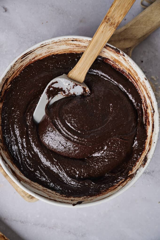 An overhead, close-up shot of thick, dark chocolate Air Fryer Slutty Brownie batter being stirred in a white ceramic bowl with a grey and bamboo spatula. The glossy batter shows a swirl texture, set against a light grey marble surface with a wooden handle visible in the background.