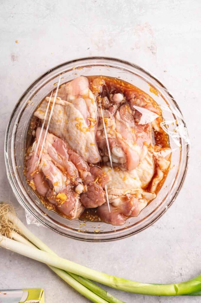 Raw chicken thighs marinating in a sticky soy-based sauce in a glass bowl covered with plastic wrap, with fresh green onions alongside, viewed from above.
