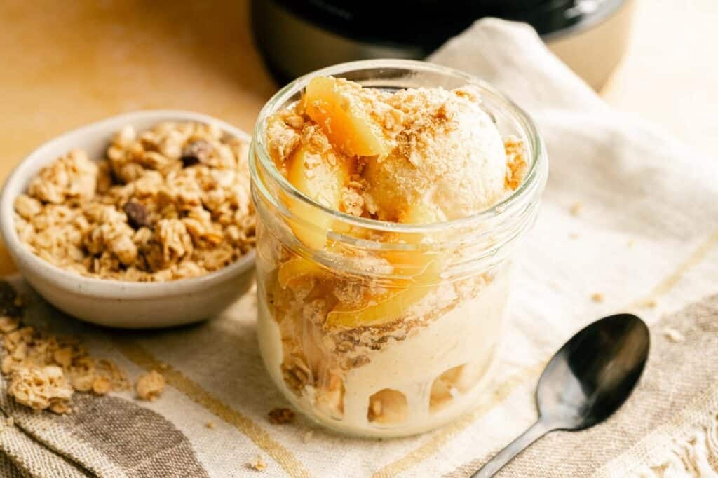 A glass jar filled with creamy peach cobbler ice cream topped with peach slices and crunchy crumble topping, placed on a light cloth beside a spoon.