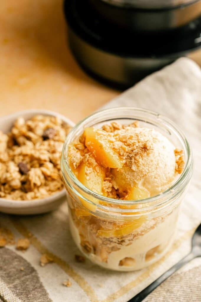 A glass jar filled with creamy peach cobbler ice cream topped with peach slices and crunchy crumble topping, placed on a light cloth beside a spoon.