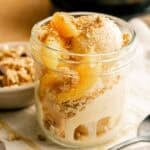 A glass jar filled with creamy peach cobbler ice cream topped with peach slices and crunchy crumble topping, placed on a light cloth beside a spoon.