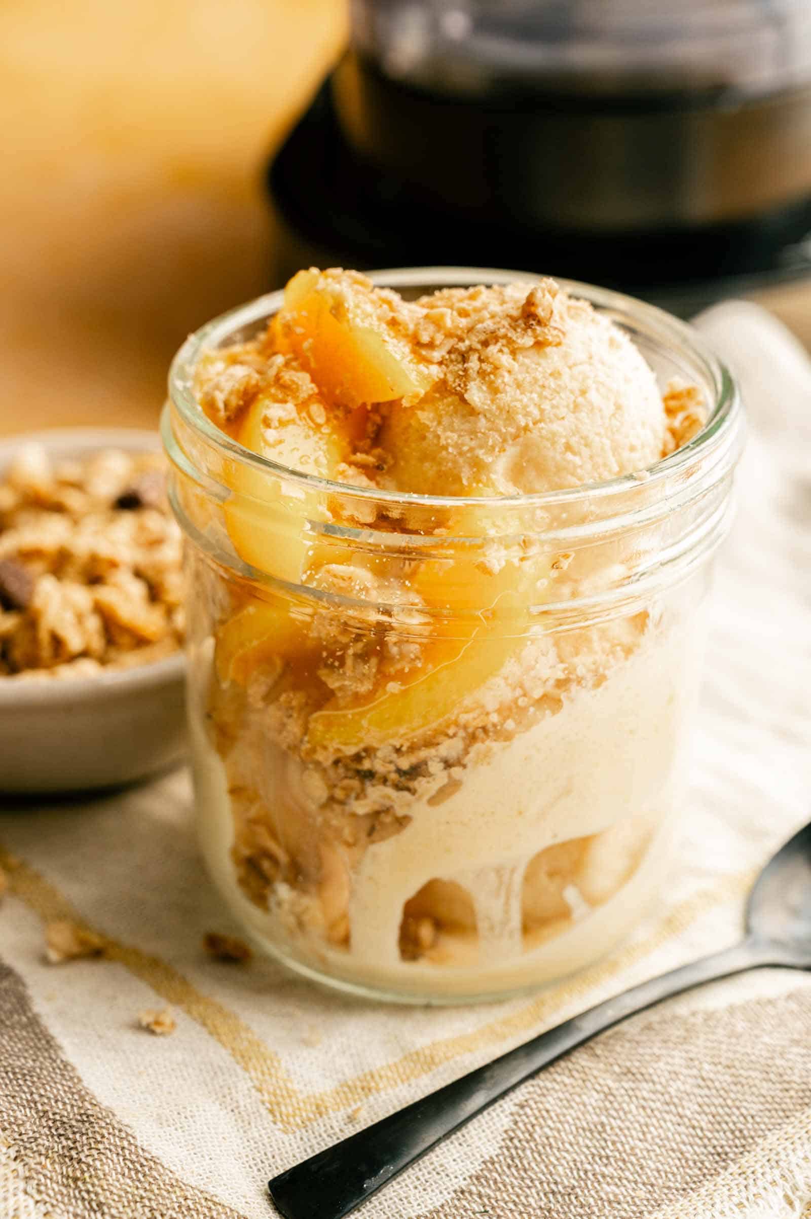 A glass jar filled with creamy peach cobbler ice cream topped with peach slices and crunchy crumble topping, placed on a light cloth beside a spoon.